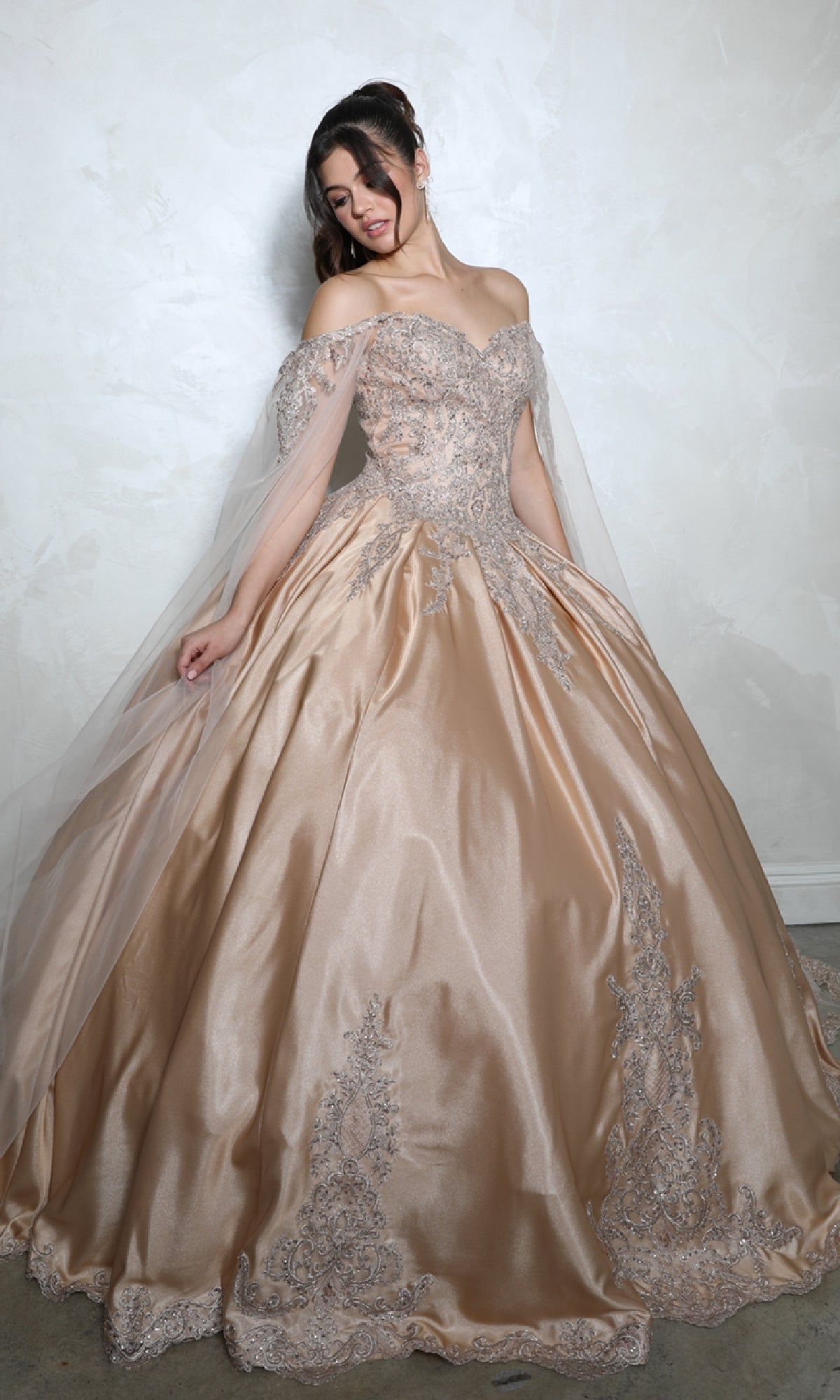 A woman in the Quinceanera Dress EK1002 By Eureka, featuring an off-the-shoulder design, intricate embroidery, and a lace-up back, stands against a light textured wall, elegantly holding the sides of her gown.